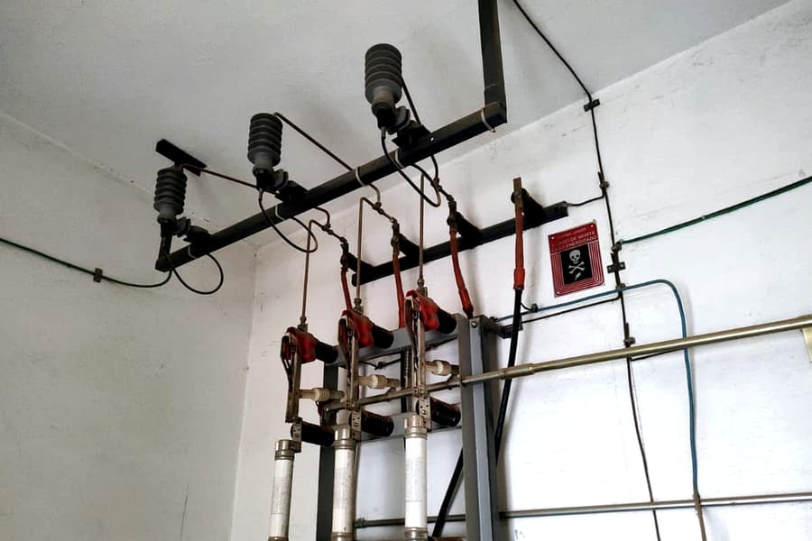 Conjunto de média tensão fixado em estrutura junto ao teto, com isoladores, chaves seccionadoras e porta-fusíveis cilíndricos aparentes, e placa de advertência de alta tensão na parede.